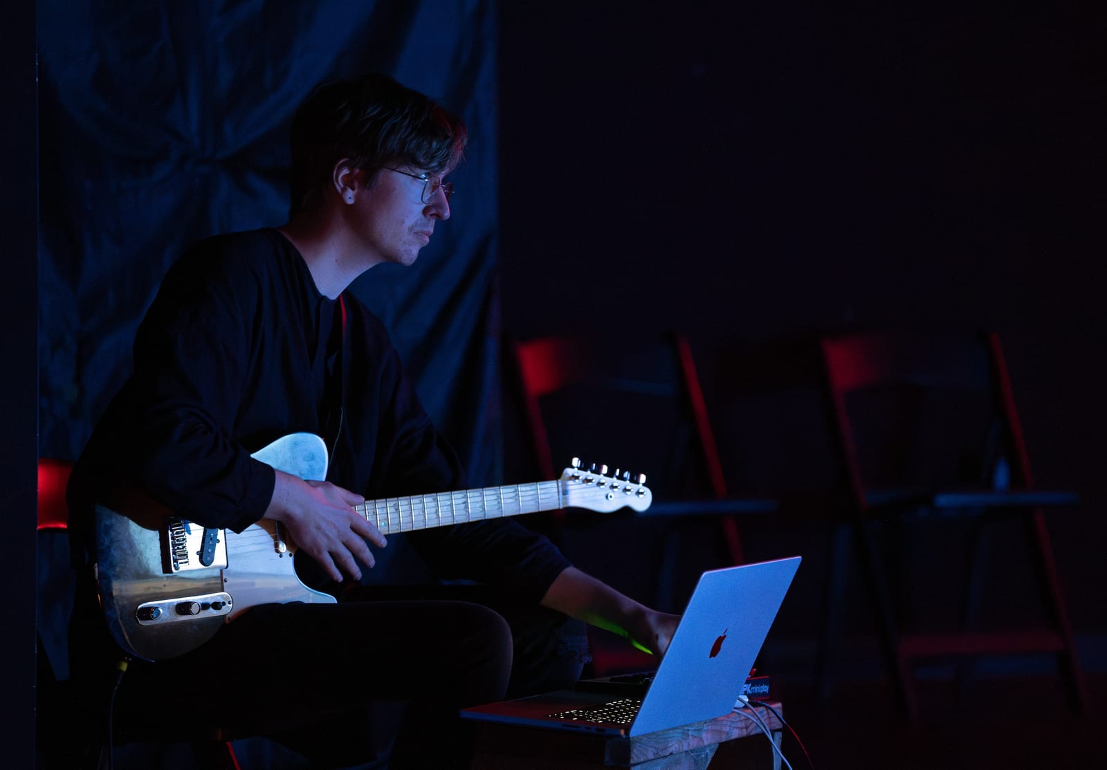 Devon Bate seated on stage with an electric guitar and a laptop, lit in blue